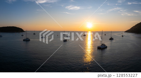 Aerial view of Naiharn Beach in Phuket, Thailand, at sunset. Sailboats and yachts rest on calm waters, framed by forested hills and a golden horizon. 128553227