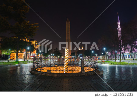 The Serpent Column in Sultanahmet Square, Istanbul, illuminated at night with the spire of the Sultan Ahmed Mosque lit in pink in the background. 128553232