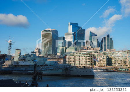 The iconic Walkie Talkie building and HMS Belfast on the River Thames, with modern skyscrapers like the Leadenhall Building under a clear blue sky. 128553251