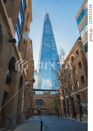 The Shard rises against a clear blue sky, framed by historic brick buildings. A cobblestone street leads to its base, with bare trees indicating winter. The Shard rises against a clear blue sky, framed by historic brick buildings. A cobblestone street leads to its base, with bare trees indicating winter. 128553338