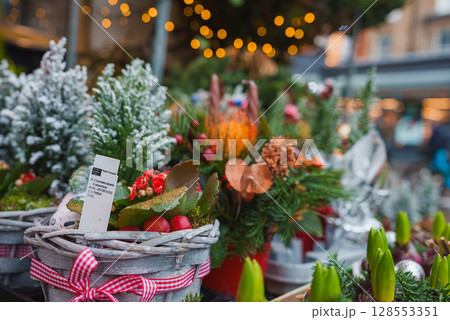 A vibrant display of potted evergreens, floral arrangements, and ornaments at a London Christmas market. Warm lights create a cozy holiday ambiance. A vibrant display of potted evergreens, floral arrangements, and ornaments at a London Christmas market. Warm lights create a cozy holiday ambiance. 128553351