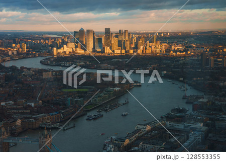 The image shows an aerial view of London's Tower Bridge with its twin towers, the River Thames, and Canary Wharf skyscrapers under a golden light. The image shows an aerial view of London's Tower Bridge with its twin towers, the River Thames, and Canary Wharf skyscrapers under a golden light. 128553355