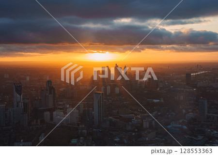 The image shows a sunset over London with Canary Wharf skyscrapers, a golden glow on the horizon, dark clouds, and the River Thames winding through the city. The image shows a sunset over London with Canary Wharf skyscrapers, a golden glow on the horizon, dark clouds, and the River Thames winding through the city. 128553363