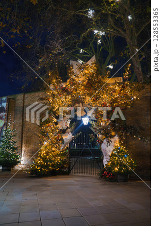 A festive archway in London adorned with twinkling lights, golden ornaments, and greenery. A lamppost and lit Christmas trees enhance the holiday scene. 128553365
