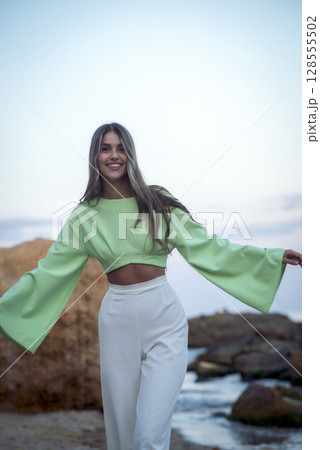 a slender young girl in white trousers stands on the seashore 128555502