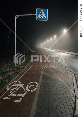 A foggy night scene with an illuminated pedestrian crossing and a row of glowing street lamps. 128556316