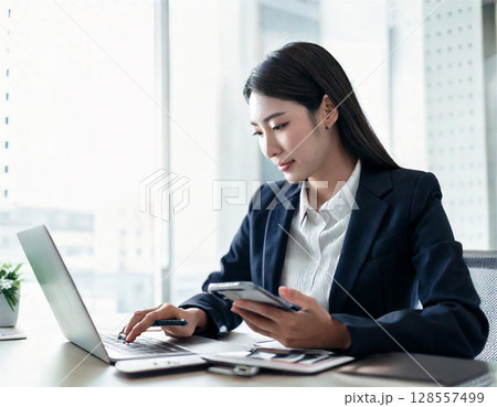 スマホとパソコンで働く女性 – リモートワーク・フリーランス・オフィス業務の現代的ビジネスシーン 128557499