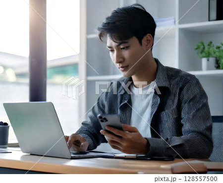 スマホとパソコンで働く男性 – リモートワーク・フリーランス・オフィス業務の現代的ビジネスシーン 128557500