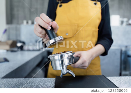 Barista Preparing Espresso with Tamper in Modern Coffee Shop Wearing Yellow Apron 128558093