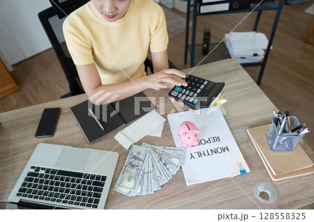 Young Woman Calculating Monthly Expenses at Home Office Desk with Calculator, Cash, and Financial Documents 128558325
