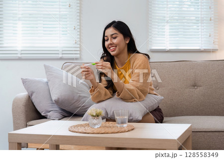 Happy Woman Excitedly Holding Positive Pregnancy Test in Cozy Living Room Setting with Natural Light Happy Woman Excitedly Holding Positive Pregnancy Test in Cozy Living Room Setting with Natural Light 128558349