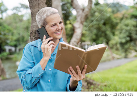 Senior Woman Enjoying a Phone Call and Reading a Book Outdoors in a Park Setting Senior Woman Enjoying a Phone Call and Reading a Book Outdoors in a Park Setting 128558419
