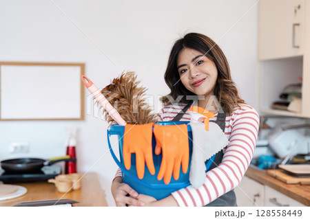 Smiling Woman Holding Cleaning Supplies and Dog in Modern Kitchen, Ready for House Cleaning Service 128558449