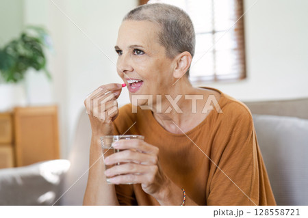 Senior Woman Taking Medication at Home, Smiling and Holding a Glass of Water in a Bright Living Room Senior Woman Taking Medication at Home, Smiling and Holding a Glass of Water in a Bright Living Room 128558721