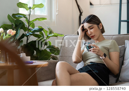 Young Woman Sitting on Couch Holding Medication Blister Pack, Looking Unwell, in a Modern Living Room with Plants Young Woman Sitting on Couch Holding Medication Blister Pack, Looking Unwell, in a Modern Living Room with Plants 128558740