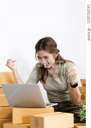 Young Woman Celebrating Success While Working on Laptop Surrounded by Cardboard Boxes at Home Office Young Woman Celebrating Success While Working on Laptop Surrounded by Cardboard Boxes at Home Office 128558741