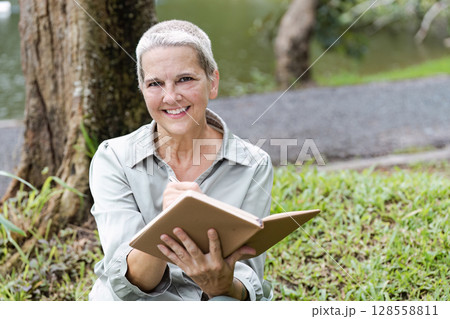 Senior Woman Writing in Journal Outdoors by a Tree Near a Lake on a Sunny Day 128558811