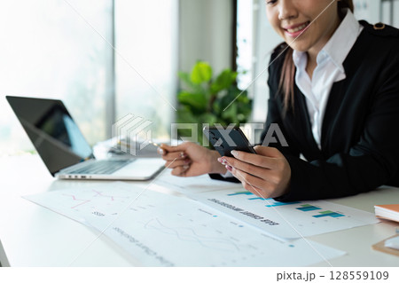 Businesswoman Analyzing Financial Data on Smartphone at Office Desk with Charts and Graphs 128559109