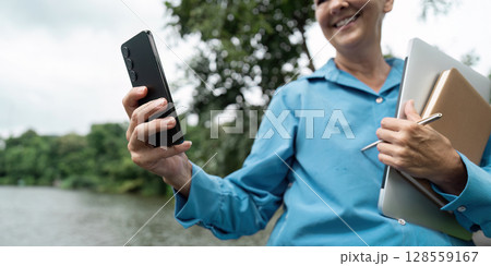 Senior Professional Enjoying Outdoor Work with Smartphone and Laptop by a Scenic Lake Senior Professional Enjoying Outdoor Work with Smartphone and Laptop by a Scenic Lake 128559167