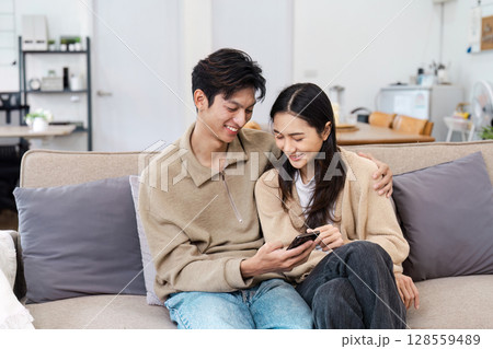 Happy Couple Relaxing on Couch at Home, Smiling and Looking at Smartphone Together in Modern Living Room Happy Couple Relaxing on Couch at Home, Smiling and Looking at Smartphone Together in Modern Living Room 128559489