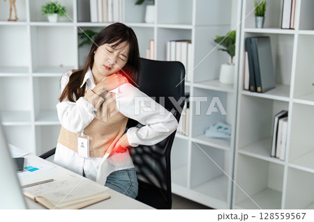 Young Woman Experiencing Shoulder Pain While Working at Desk in Modern Office Setting 128559567