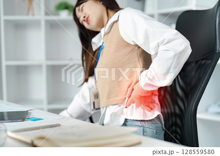 Young Woman Experiencing Severe Back Pain While Working at Desk in Modern Office Setting 128559580
