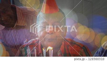 Image of glowing light over portrait of happy senior man wearing party hat blowing candles 128560310