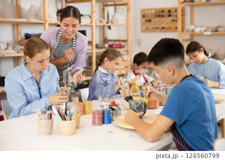 Children paint ceramic work-pieces at table with teacher in art studio 128560799