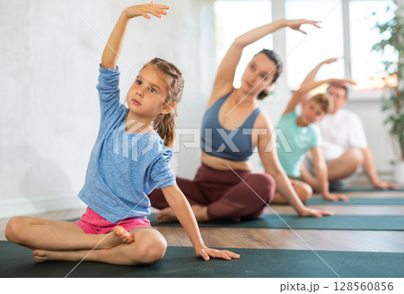Pleasant little girl practicing half lotus pose of yoga one hand raised with her family 128560856