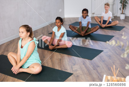 In sport studio with small group of friends, smiling girl performs butterfly pose, Baddha Konasana In sport studio with small group of friends, smiling girl performs butterfly pose, Baddha Konasana 128560898