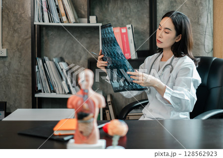 Female doctor examines X-ray in a contemporary office setting, surrounded by medical books and anatomical models. Female doctor examines X-ray in a contemporary office setting, surrounded by medical books and anatomical models. 128562923