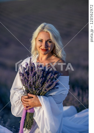 Blonde woman poses in lavender field at sunset. Happy woman in white dress holds lavender bouquet. Aromatherapy concept, lavender oil, photo session in lavender 128562972
