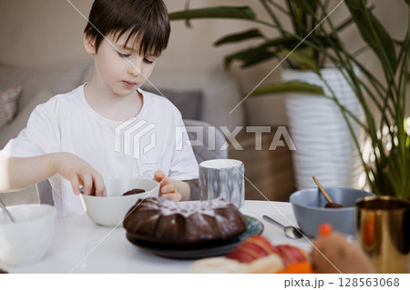Young boy having breakfast at home, eating cake at table with mugs and bowls, cozy morning 128563068