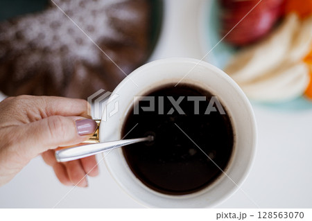 Close-up of a hand holding a coffee cup with cake and fruit on a bright breakfast table 128563070
