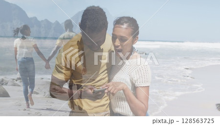 Composite of happy african american couple using smartphone, and walking holding hands on beach 128563725