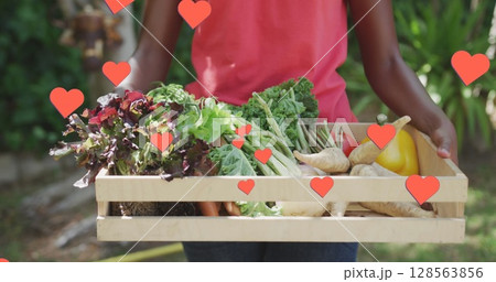 Image of hearts over happy african american woman holding box with vegetables 128563856