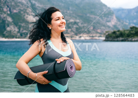 Smiling woman holding yoga mat by the sea, enjoying nature and healthy coastal lifestyle 128564483
