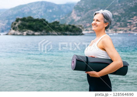 Happy senior woman with yoga mat on beach, embracing fitness and aging gracefully outdoors Happy senior woman with yoga mat on beach, embracing fitness and aging gracefully outdoors 128564690