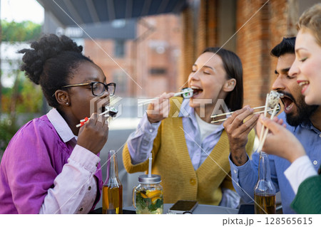 A diverse group of friends enjoys sushi outdoors, a moment of shared joy and culinary delight. A diverse group of friends enjoys sushi outdoors, a moment of shared joy and culinary delight. 128565615