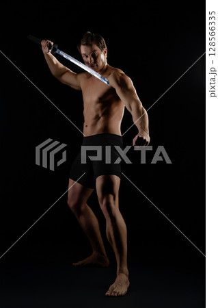 Strong man performing a dynamic sword technique indoors in low light setting 128566335