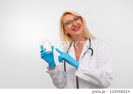 Middle aged blonde therapist in white coat, gloves, and stethoscope holding a bottle of vitamins. Isolated on white background with copy space Middle aged blonde therapist in white coat, gloves, and stethoscope holding a bottle of vitamins. Isolated on white background with copy space 128566824