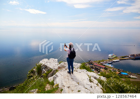 A female traveler stands on a rock near the port of Baikal and photographs Lake Baikal  128567464