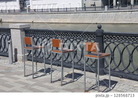 A row of chairs with wooden seats covered with snow near a metal fence on the canal embankment. 128568713