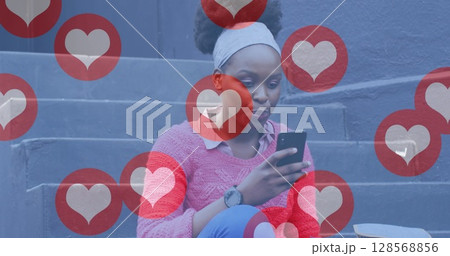 Image of social media heart icons over happy african american woman in street using smartphone 128568856