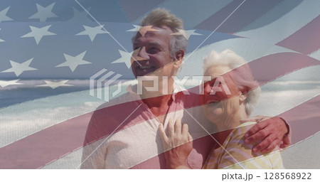 Image of happy senior caucasian couple embracing on beach over flag of united states of america 128568922