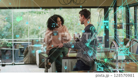Image of network of media icons over happy biracial couple having coffee talking at home Image of network of media icons over happy biracial couple having coffee talking at home 128570981