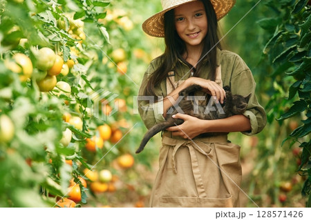 Holding cute kitten in hands. Little girl is in the garden with tomatoes 128571426
