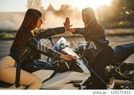 Giving high five to each other. Two women on motorcycles are together outdoors 128571918
