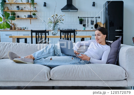 Portrait of young woman lying on sofa typing on laptop, in home interior 128572079