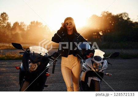 Holding protective helmet in hand. Woman is with motorcycle outdoors 128572133
